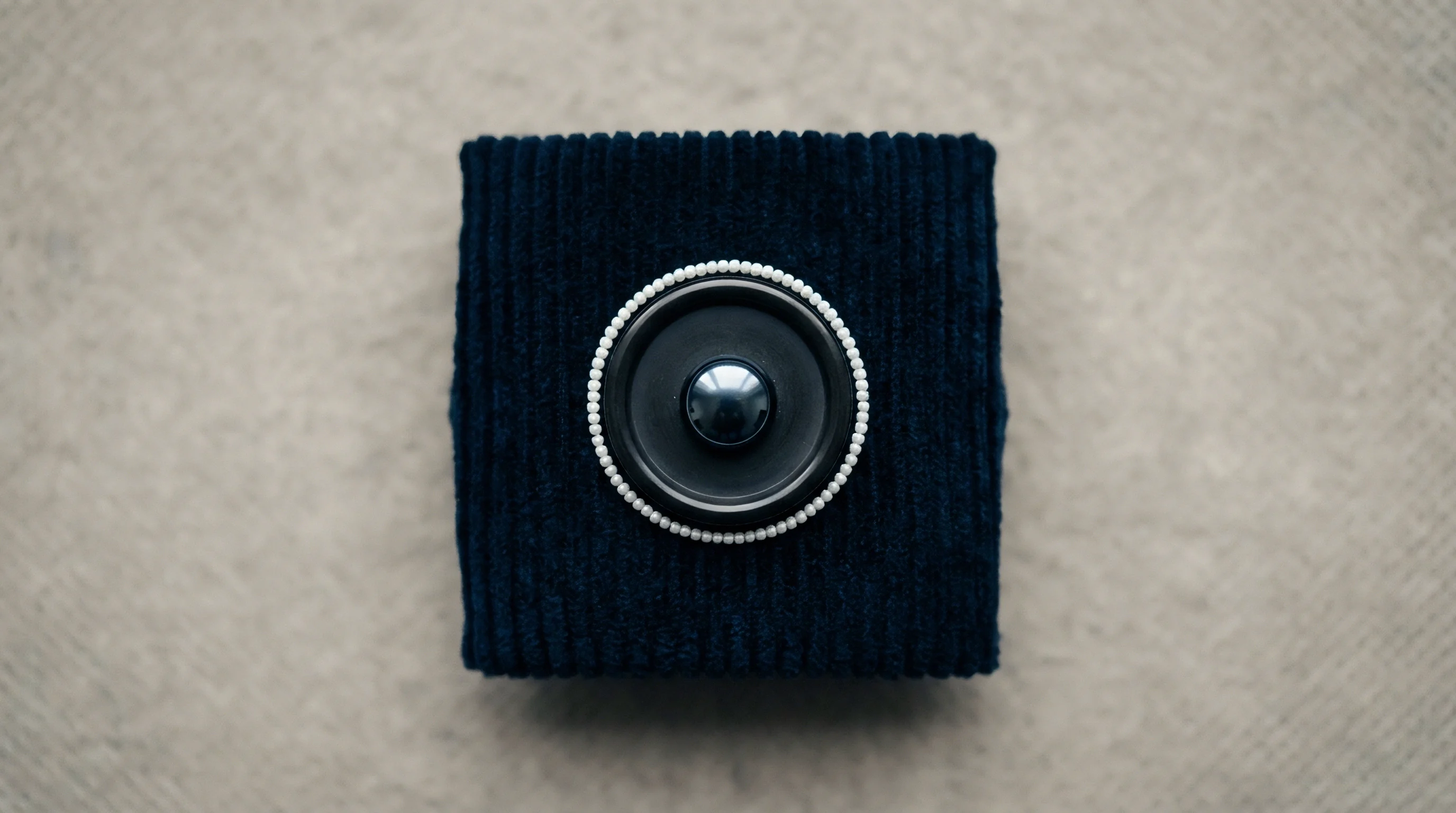 A navy blue corduroy fabric-covered square object features a central dark speaker driver surrounded by a beaded trim.