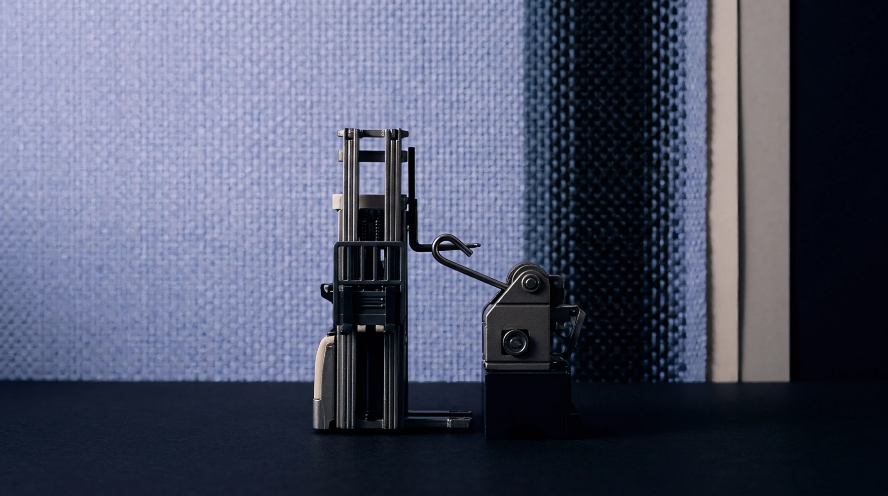 Miniature industrial machinery stands on a dark surface against a textured blue woven fabric backdrop with a vertical dark blue stripe.
